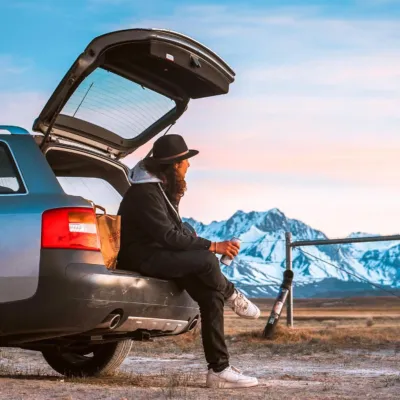 Person relaxing under an open car trunk safely held up by reliable xterra hatch struts, facing snowy mountains.