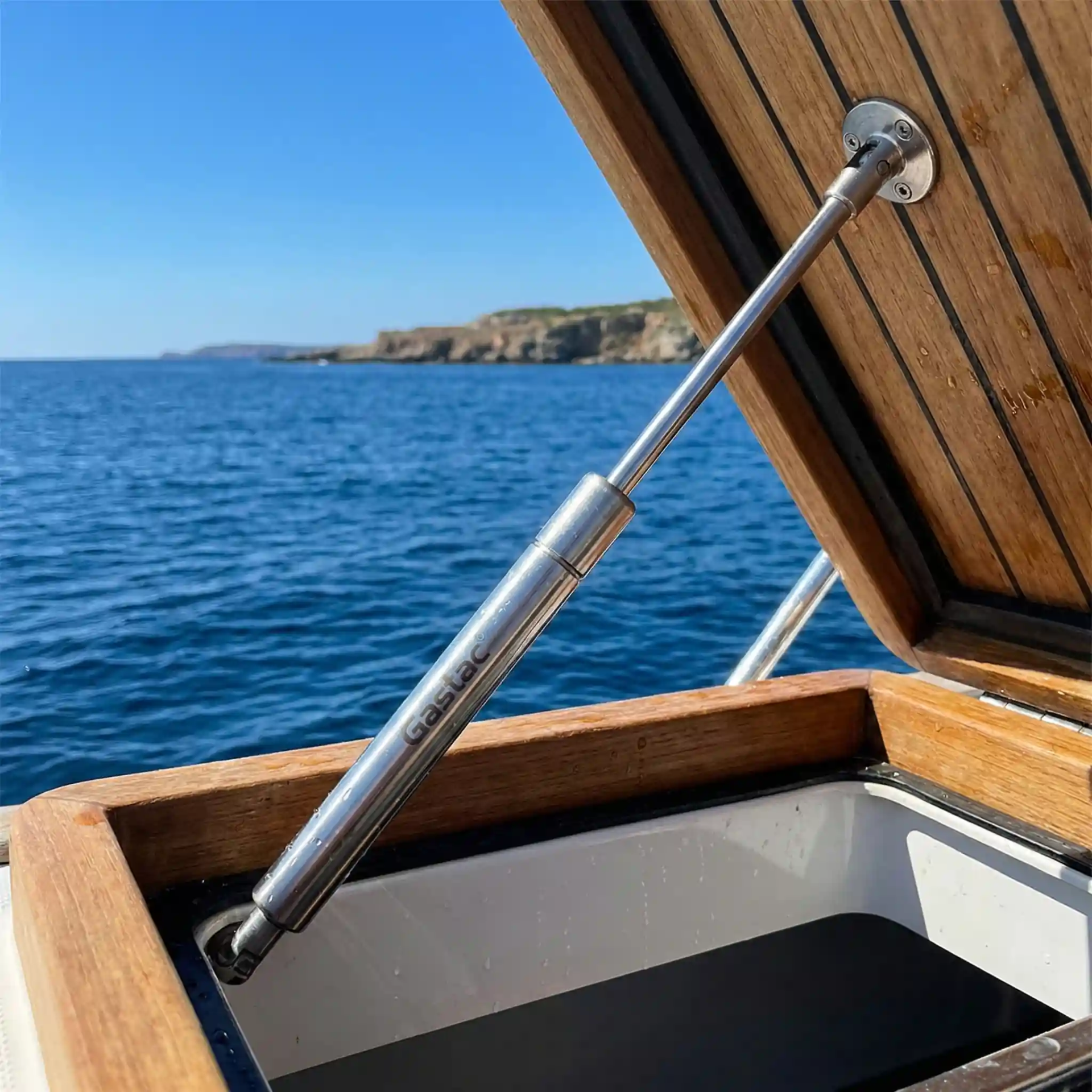 A close-up view of a Gastac branded stainless steel marine gas strut supporting an open teak wood boat hatch, with a blue ocean and coastline in the background.