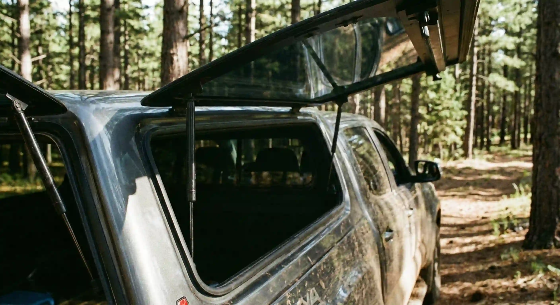 truck-camper-shell-side-door-open-with-gas-struts-forest Side view of a truck camper shell door held open by gas struts in a forest.