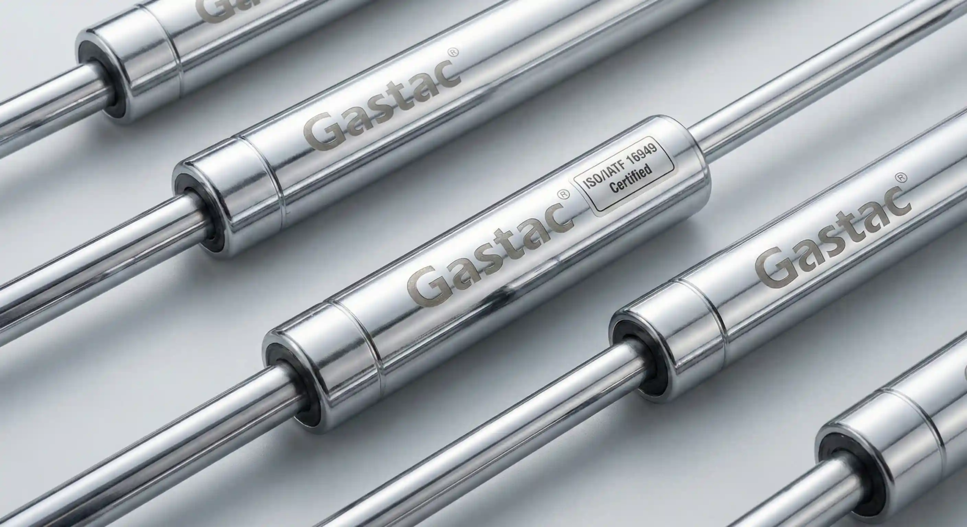 A premium studio photograph of multiple polished metallic Gastac gas springs arranged neatly diagonally on a seamless light gray background. The "Gastac" brand logo and an "ISO/IATF 16949 Certified" label are clearly engraved on the cylinder bodies.